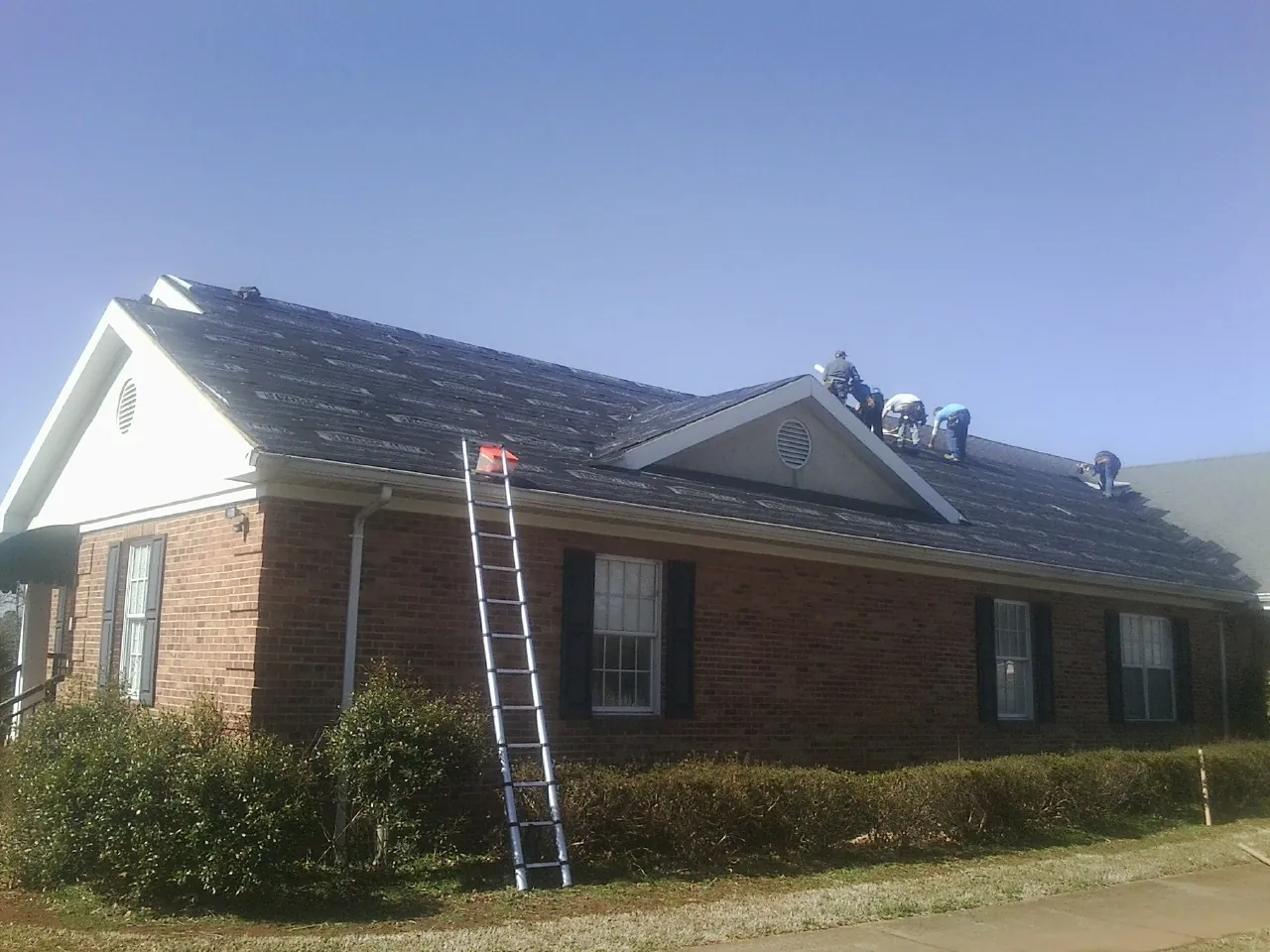 Crew tearing shingles off a brick ranch roof with a ladder set — Metal Roof Repair work by our crew in East Spencer