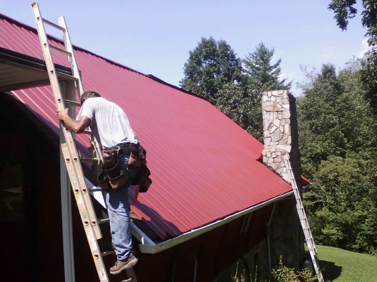 Roofer on a ladder beside a red metal roof and stone chimney — Soffit & Fascia Repair by Roofing Brothers in East Spencer